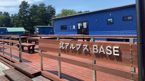 A wooden bench with the text "北ルブス BASE" and a blue building in the background. - Kita-alps BASE Onsenkyo Camp (Omachi)
