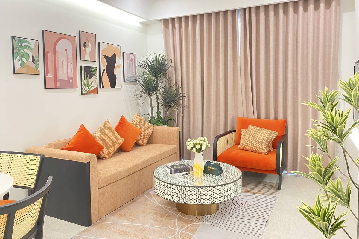 Een moderne woonkamer met een bank, fauteuil en een rond bijzettafeltje. Er hangen kunstwerken in lijsten aan de muur, een potplant en een gordijn.