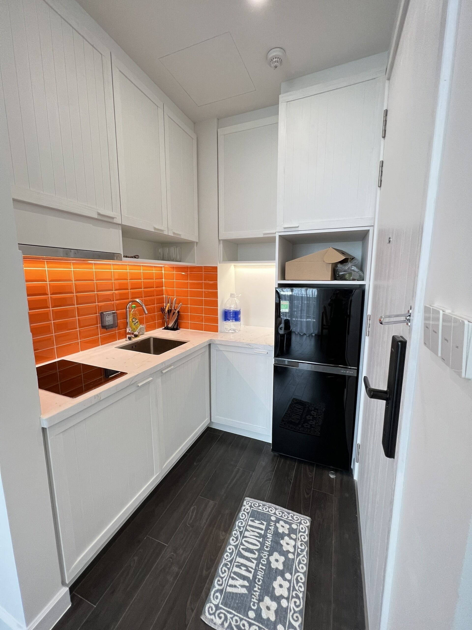 Una cocina compacta con armarios blancos, un refrigerador negro y un salpicadero de azulejos naranja.
