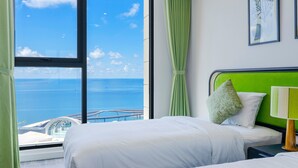Une chambre d’hôtel dotée d’une grande fenêtre donnant sur la mer et un pont. La chambre comprend deux lits avec du linge de lit blanc et des rideaux verts.
