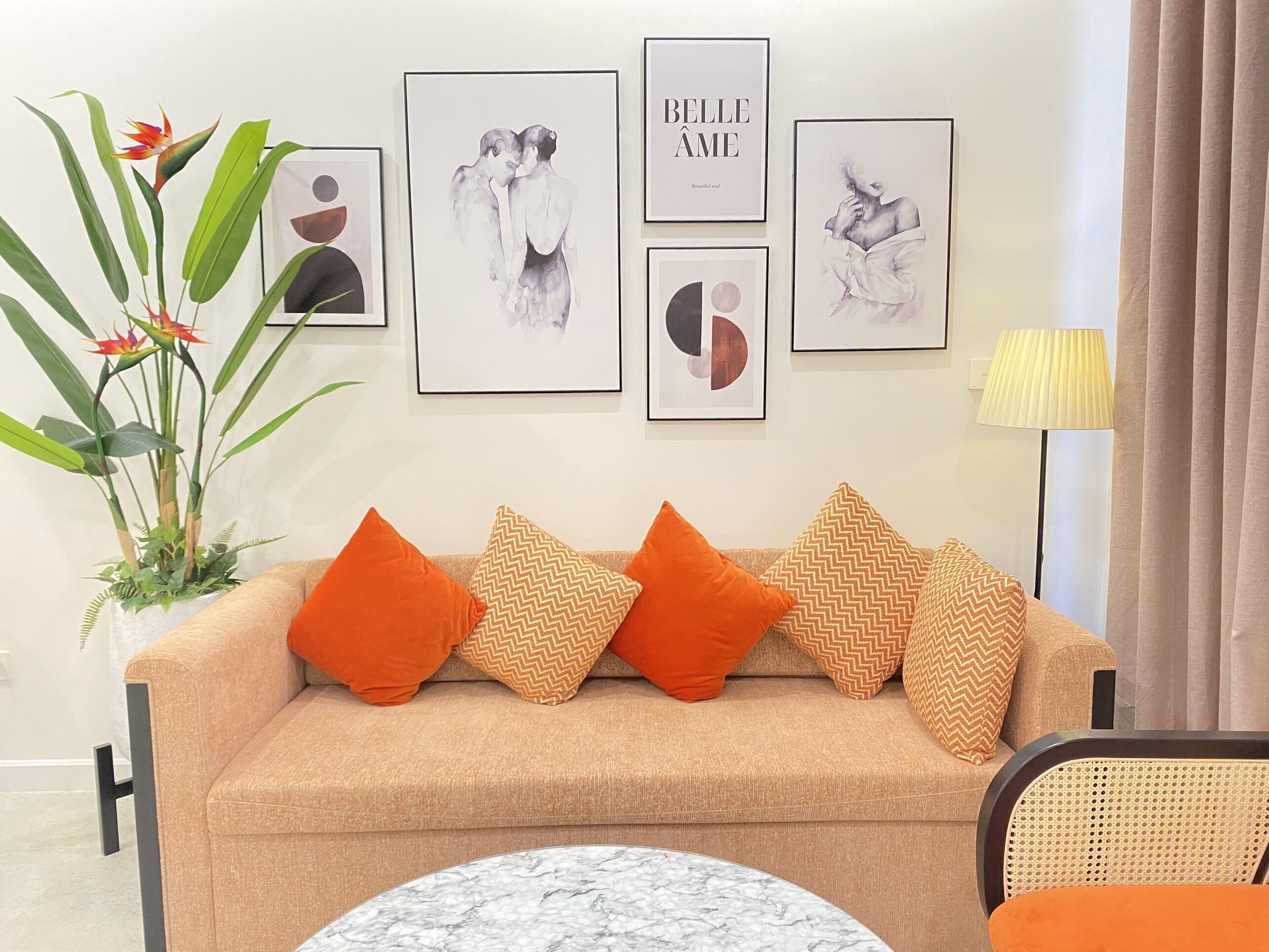 Uma sala de estar com sofá bege, almofadas laranja e estampadas, uma mesa de centro de mármore redonda e obras de arte nas paredes, incluindo um quadro com um casal e outro com a inscrição "BELLE AME".