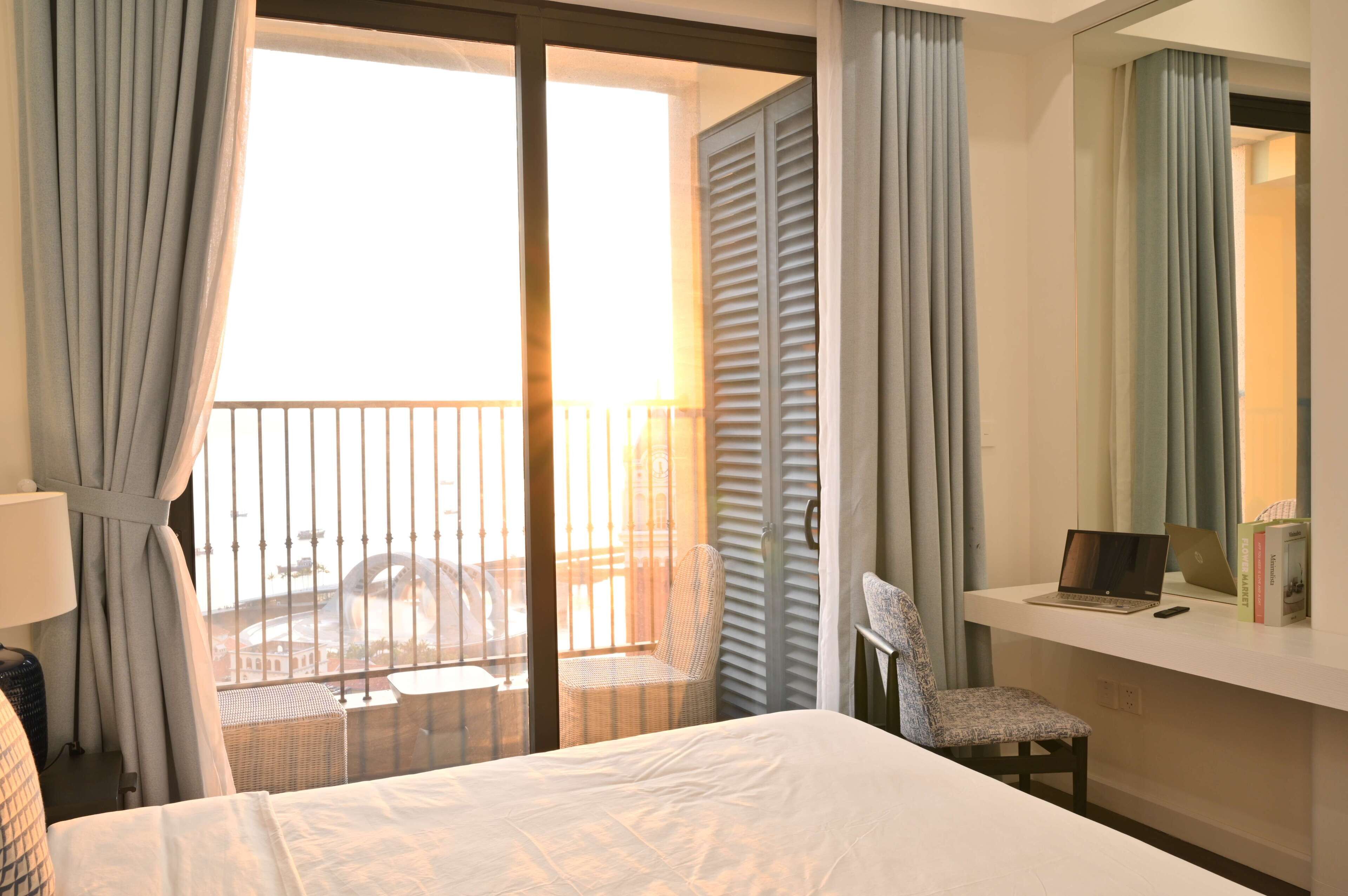 A hotel room with a bed, a desk with a laptop, a chair, and a balcony with a view.