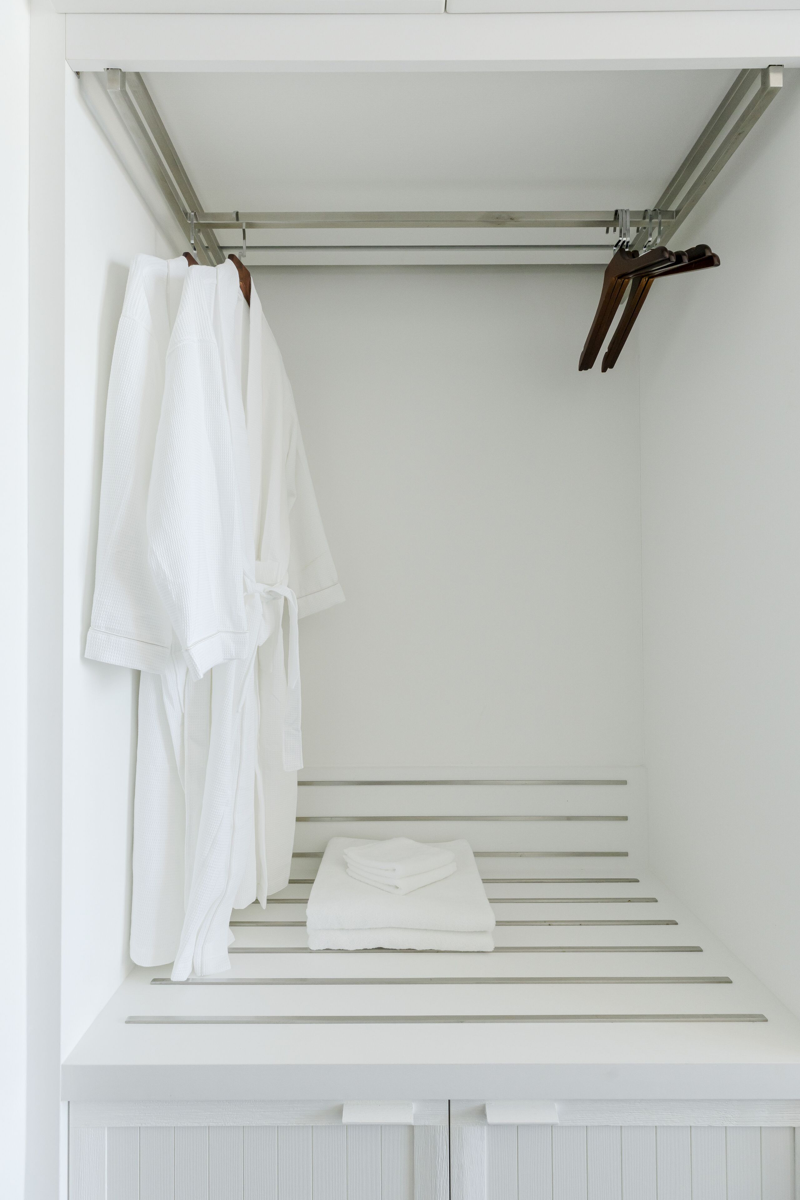 A white walk-in closet with a metal rod for hanging clothes, a folded white towel on the lower shelf, and a pair of brown shoes on the rod.