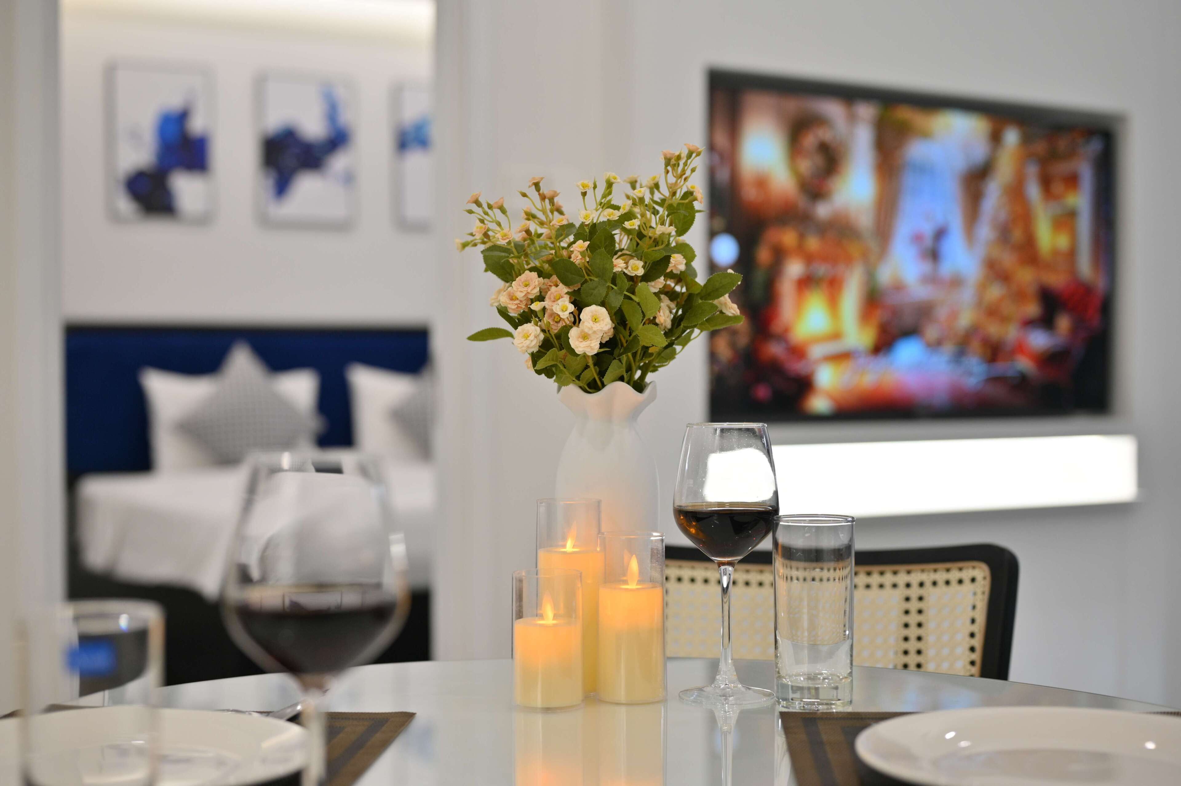 A dining table with candles, wine glasses, and a vase of flowers. A bed and a television are visible in the background.