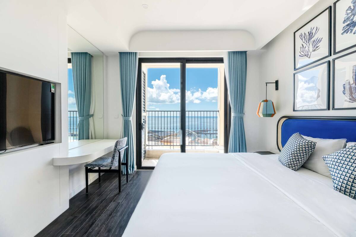 Een hotelkamer met een bed, een bureau, een stoel en een balkon met uitzicht op zee.