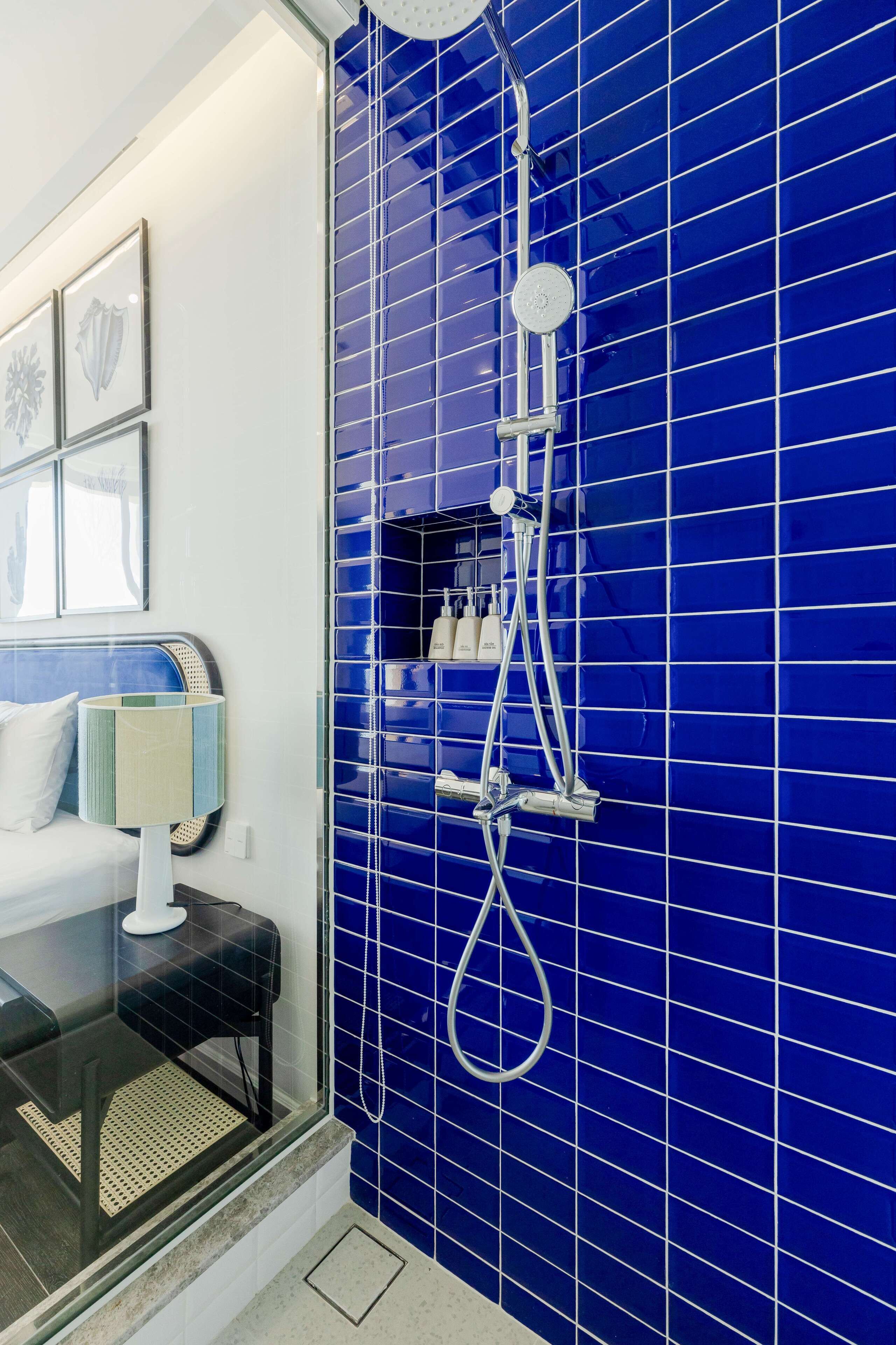 Casa de banho com paredes azulejadas em azul, cabine de duche em vidro e uma cama com mesa de cabeceira.