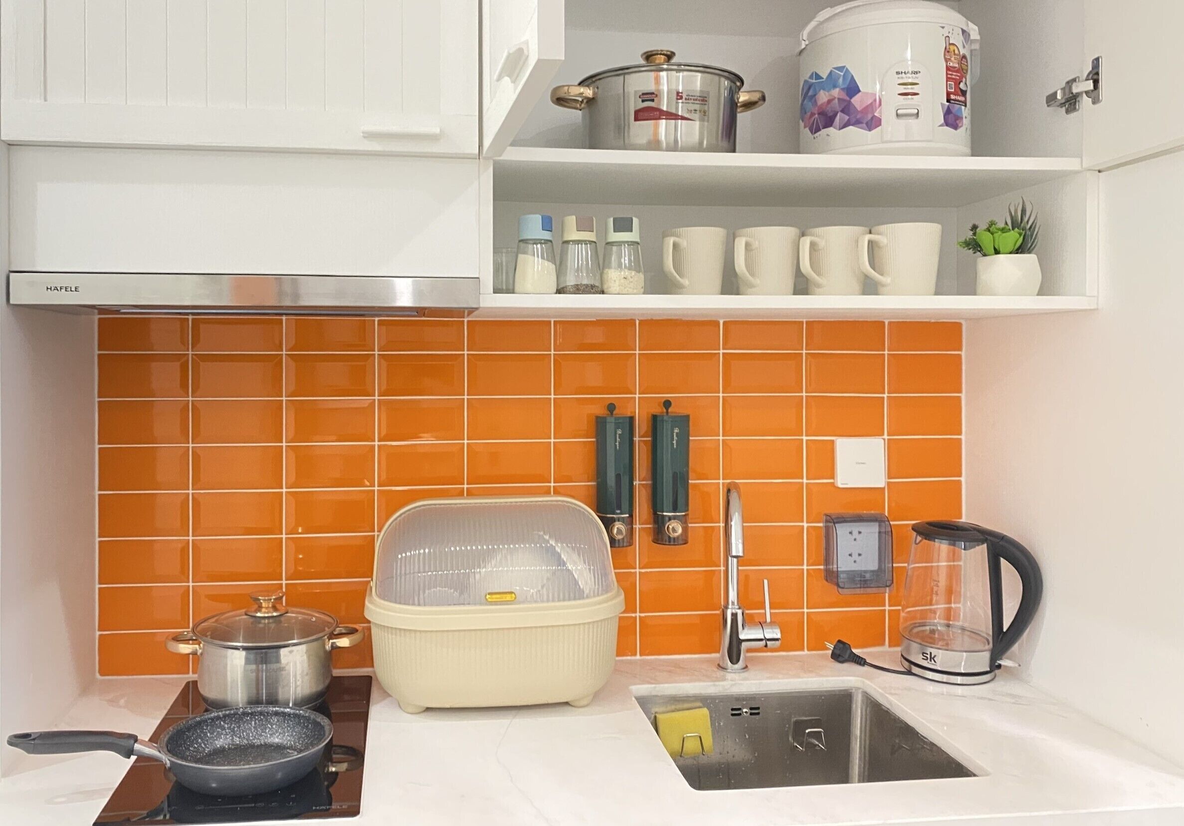 A kitchen with orange tiles, a stove, a sink, a kettle, and various kitchen items on shelves.
