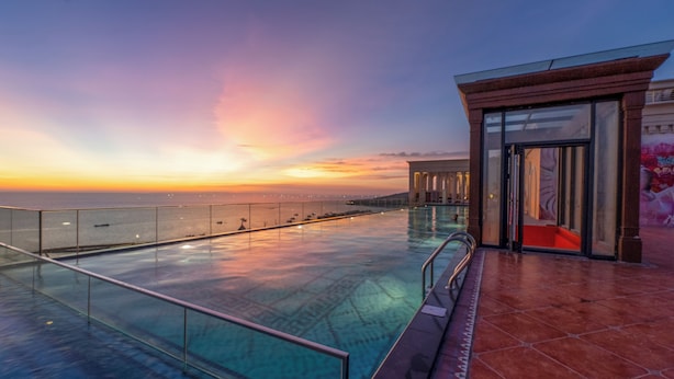 Une piscine à débordement offrant une vue imprenable sur le coucher de soleil sur l’eau, un espace salon couvert et une entrée fermée par des parois de verre.