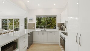 A modern kitchen with white cabinetry, a built-in dishwasher, and a large window overlooking greenery. - Villa Luna (Avoca Beach)