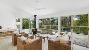 A spacious living room with wicker furniture, a fireplace, and large windows offering a view of the outdoors. - Villa Luna (Avoca Beach)