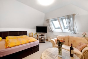 Un dormitorio con cama, televisor, sofá, una mesa con botella y vaso, y ventana con cortinas.