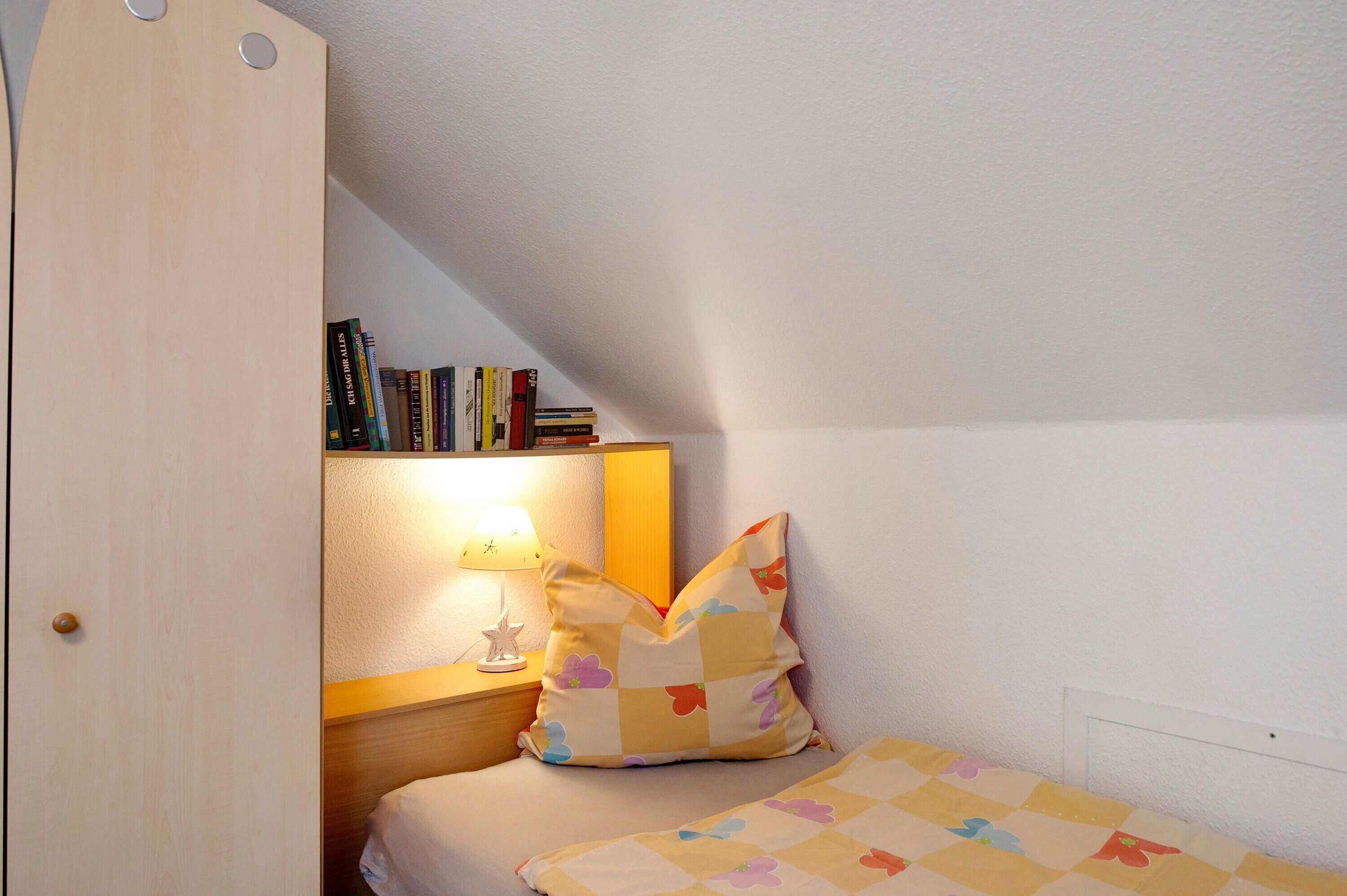 Un dormitorio con techo inclinado, una mesita de noche con lámpara, una pila de libros y una cama con colcha.