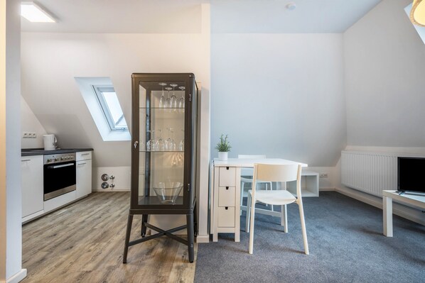 A compact kitchen with a built-in oven, a dining area with a white table and chair, and a black cabinet with glassware.