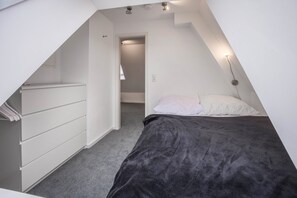 A bedroom with a sloped ceiling, a bed with a dark blanket, a white dresser, and a wall-mounted light fixture.