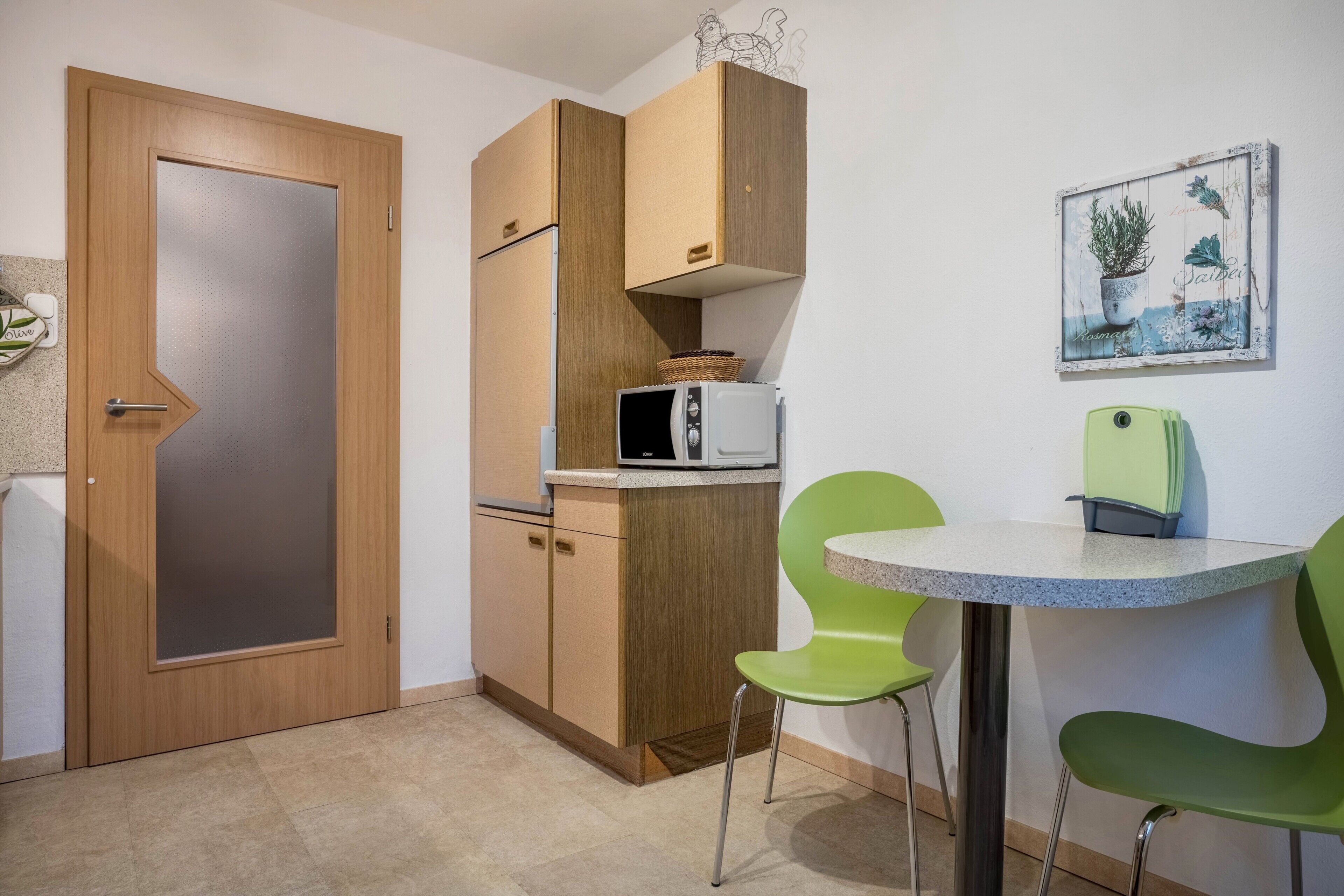 A compact kitchen with a microwave, a small refrigerator, wooden cabinets, a green chair, and a small table.