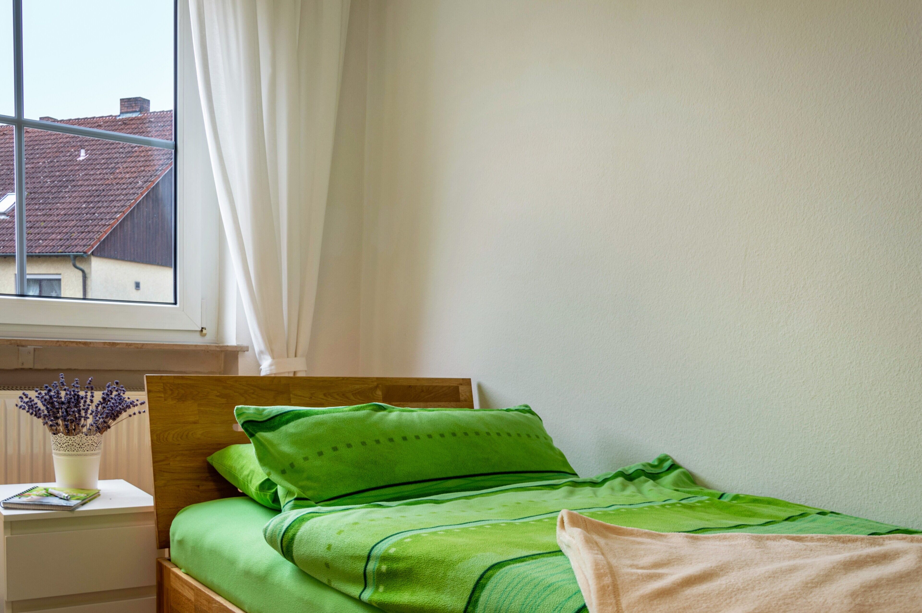 Une chambre à coucher avec une tête de lit en bois, une couvre-lit verte, une table de chevet blanche ornée d’un vase de fleurs et une fenêtre donnant sur des immeubles.