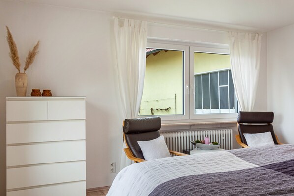 Un dormitorio con cama, dos sillones, un aparador blanco y una ventana con cortinas.