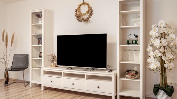 Ein Wohnzimmer mit einem Flachbildfernseher, weißen Regaleinheiten, einem dekorativen Kranz und einer Topf-Orchidee.