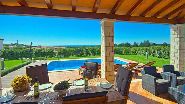 A covered outdoor dining area with a pool view, wooden furniture, and a clear sky.