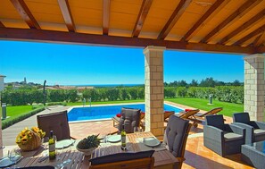 Un espace extérieur abrité, aménagé pour les repas, avec vue sur la piscine, du mobilier en bois et un ciel dégagé.