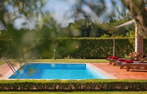Une piscine rectangulaire dont le bord est recouvert de carreaux, entourée d’une terrasse dallée. On y trouve des chaises longues avec des coussins rouges et, au fond, une structure en forme de pergola.