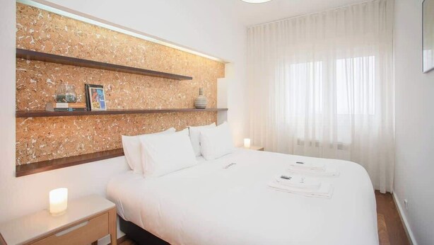 Una habitación de hotel moderna con una cama grande, cabecera de madera, mesita de noche y ventana con cortinas traslúcidas.