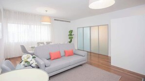 Una sala de estar moderna con un sofá gris, una mesa de centro blanca y un gran ventanal con cortinas traslúcidas.