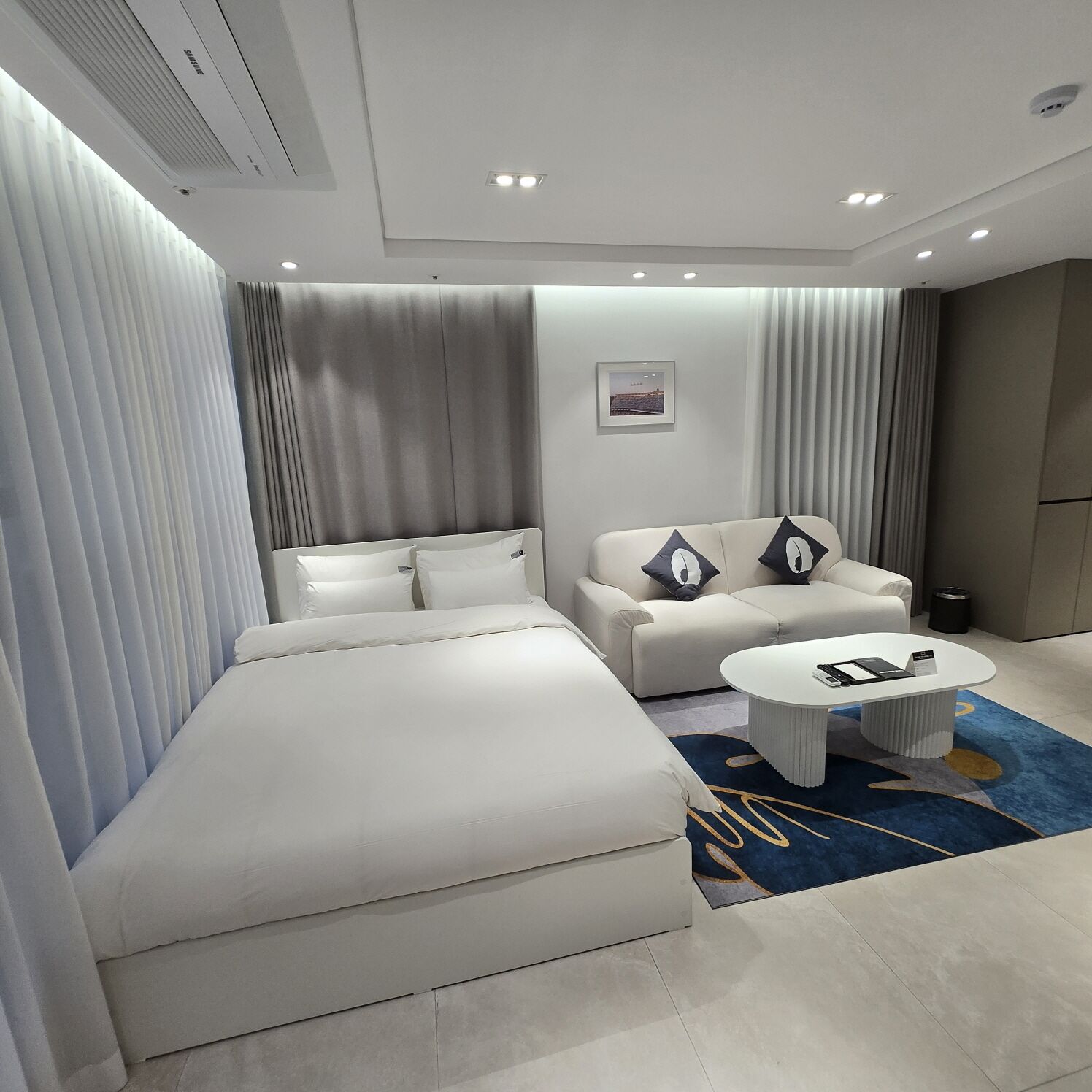 Quarto de hotel moderno com uma cama grande, um sofá, uma mesa de centro e vista para a paisagem urbana pela janela.