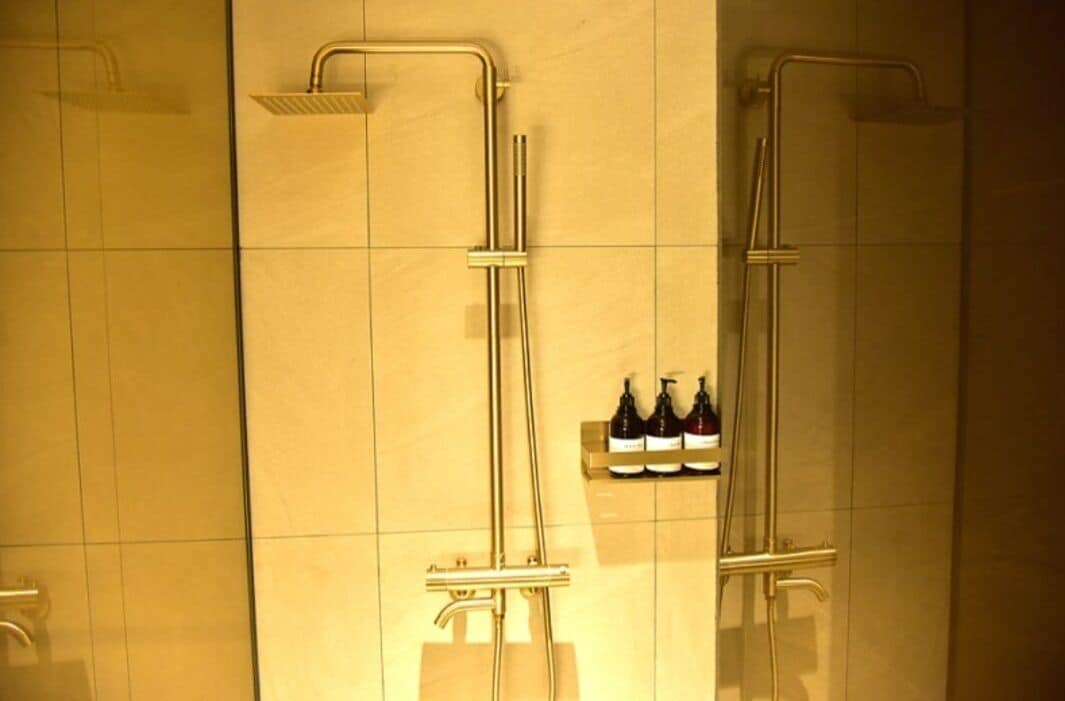 A bathroom with two showerheads, a hand dryer, and a shelf with bottles.