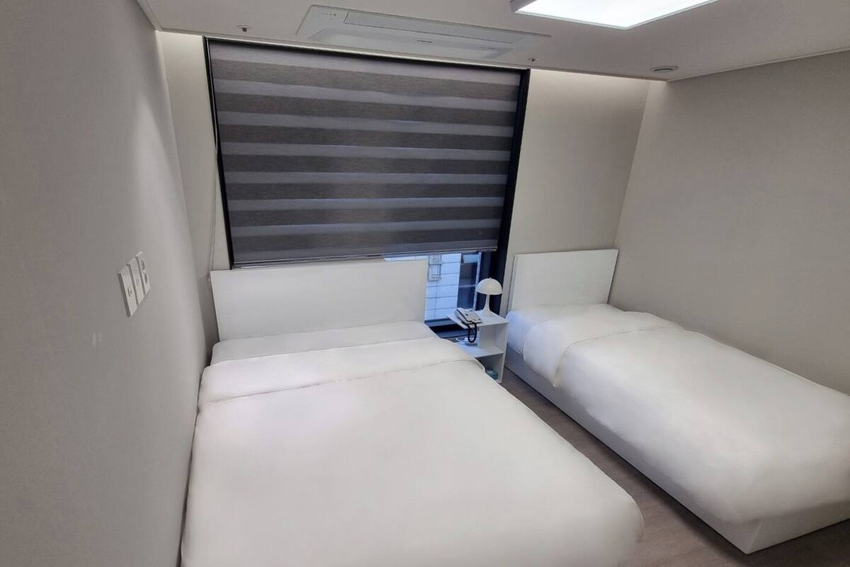 Een moderne, compacte kamer met twee eenpersoonsbedden, een klein nachtkastje met lamp en een raam met horizontale zonwering.