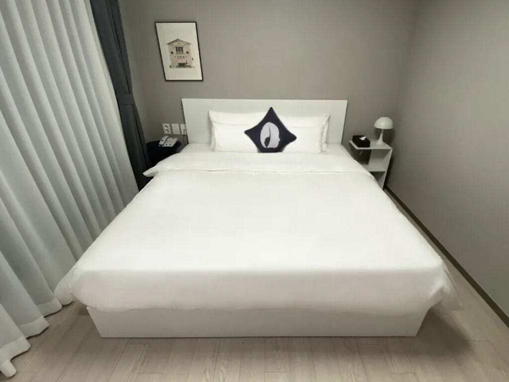 Una cama bien hecha con edredón blanco y un almohadón de diseño único. Hay una mesita de noche con una lámpara y un teléfono. La habitación tiene paredes de tonos claros y una ventana con persianas.