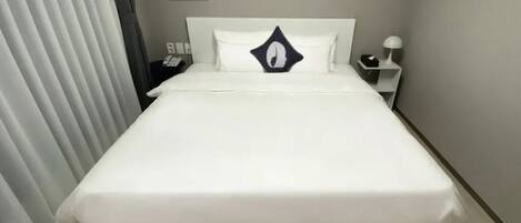 Una cama bien hecha con edredón blanco y un almohadón de diseño único. Hay una mesita de noche con una lámpara y un teléfono. La habitación tiene paredes de tonos claros y una ventana con persianas.