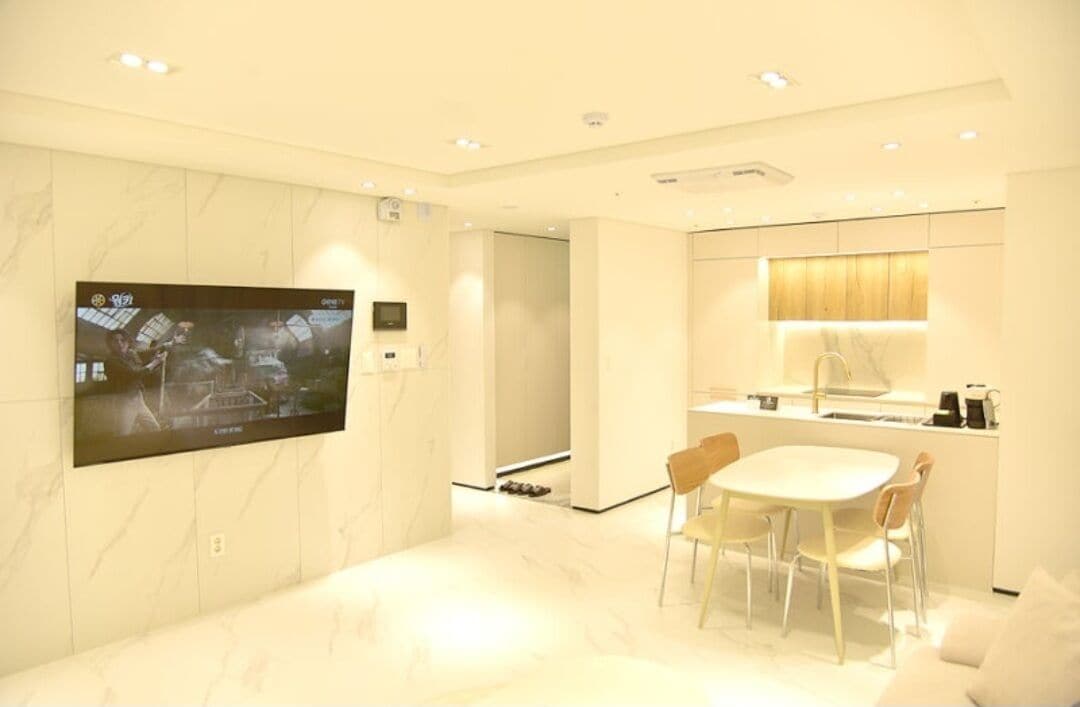 Una sala de estar moderna con un televisor de pantalla plana montado en la pared, un área de comedor con una mesa y sillas, y una cocina con un lavaplatos y alacenas.