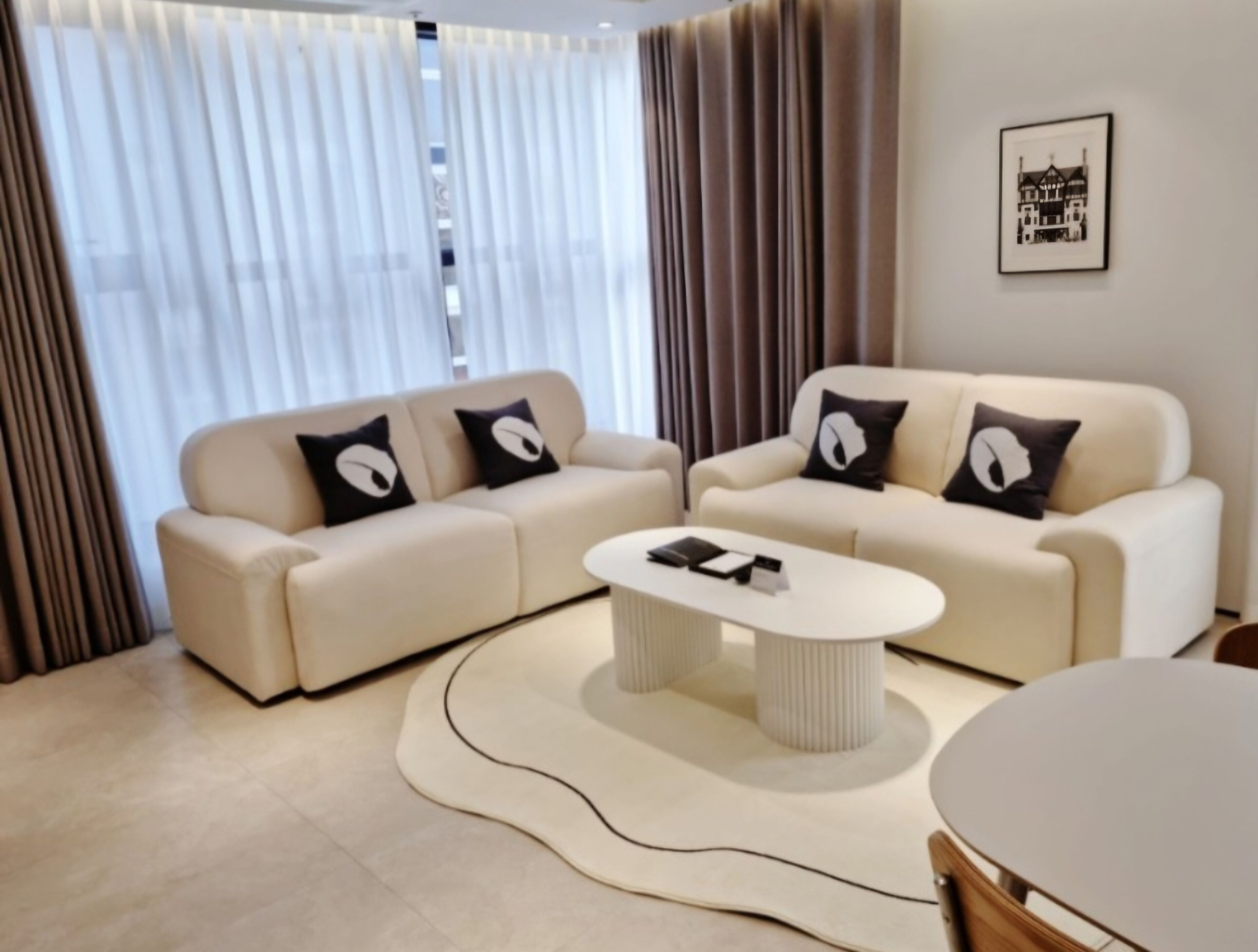 A modern living room with a beige sofa set, a round coffee table, and a large window with sheer curtains.