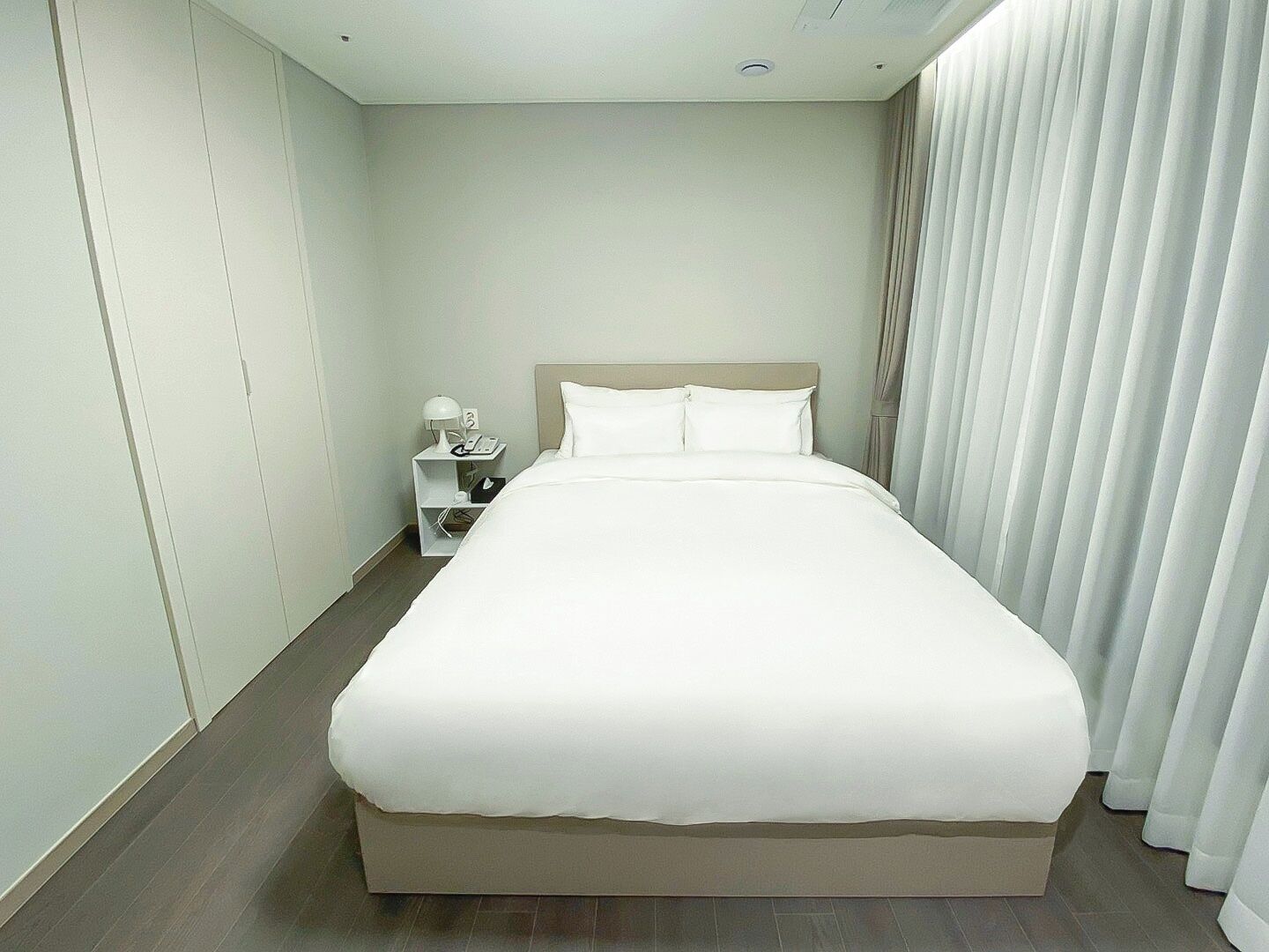 A neatly arranged hotel room with a large bed, a bedside table with a lamp, and a window with curtains.