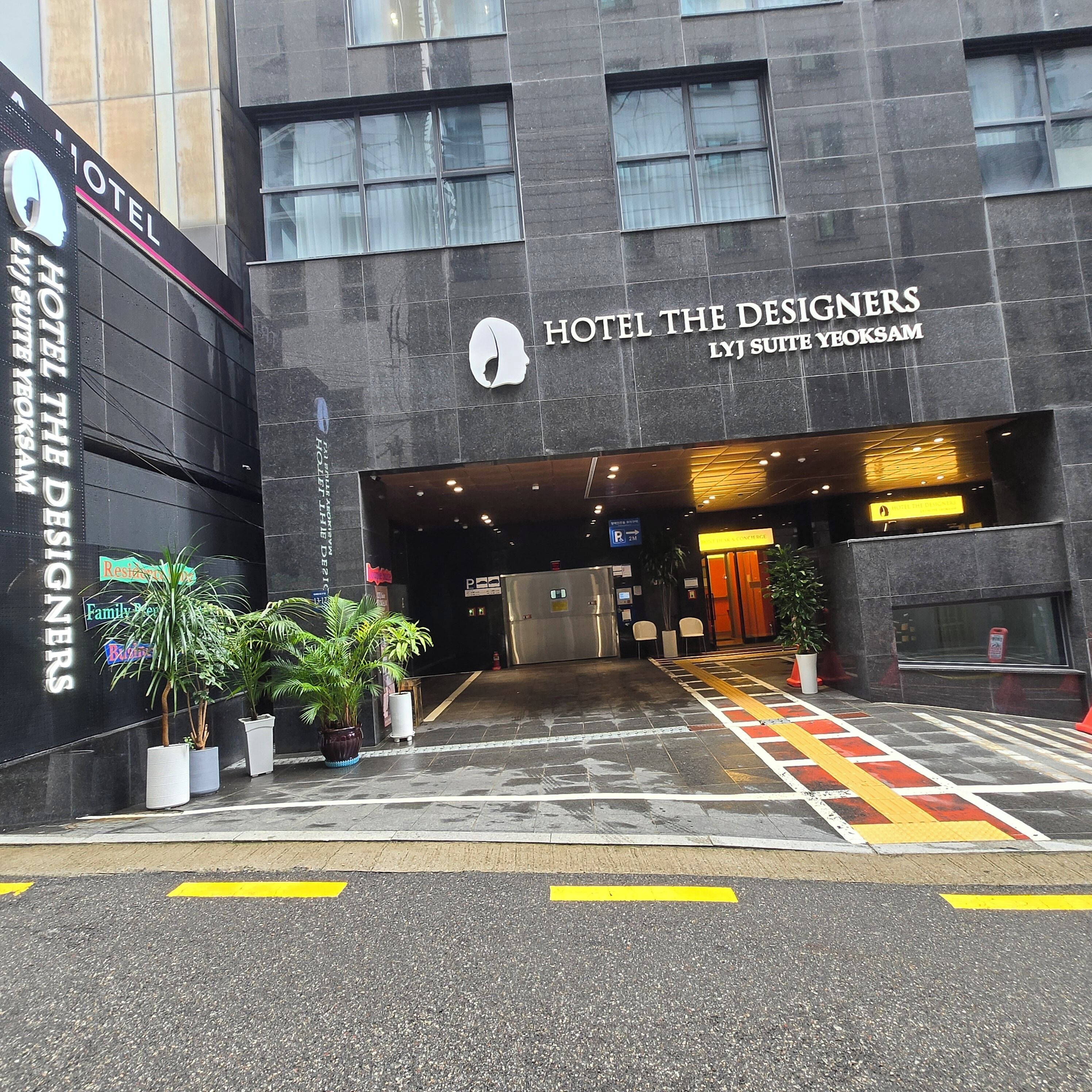Der Eingang zum Hotel The Designers, mit einer modernen Fassade, Blumentöpfen und einem deutlichen Weg zum Eingang.