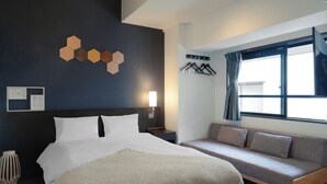 Moderni hotellihuone, jossa on suuri sänky, sohva, seinälle kiinnitetty naulakko ja ikkuna, jossa on verhot.