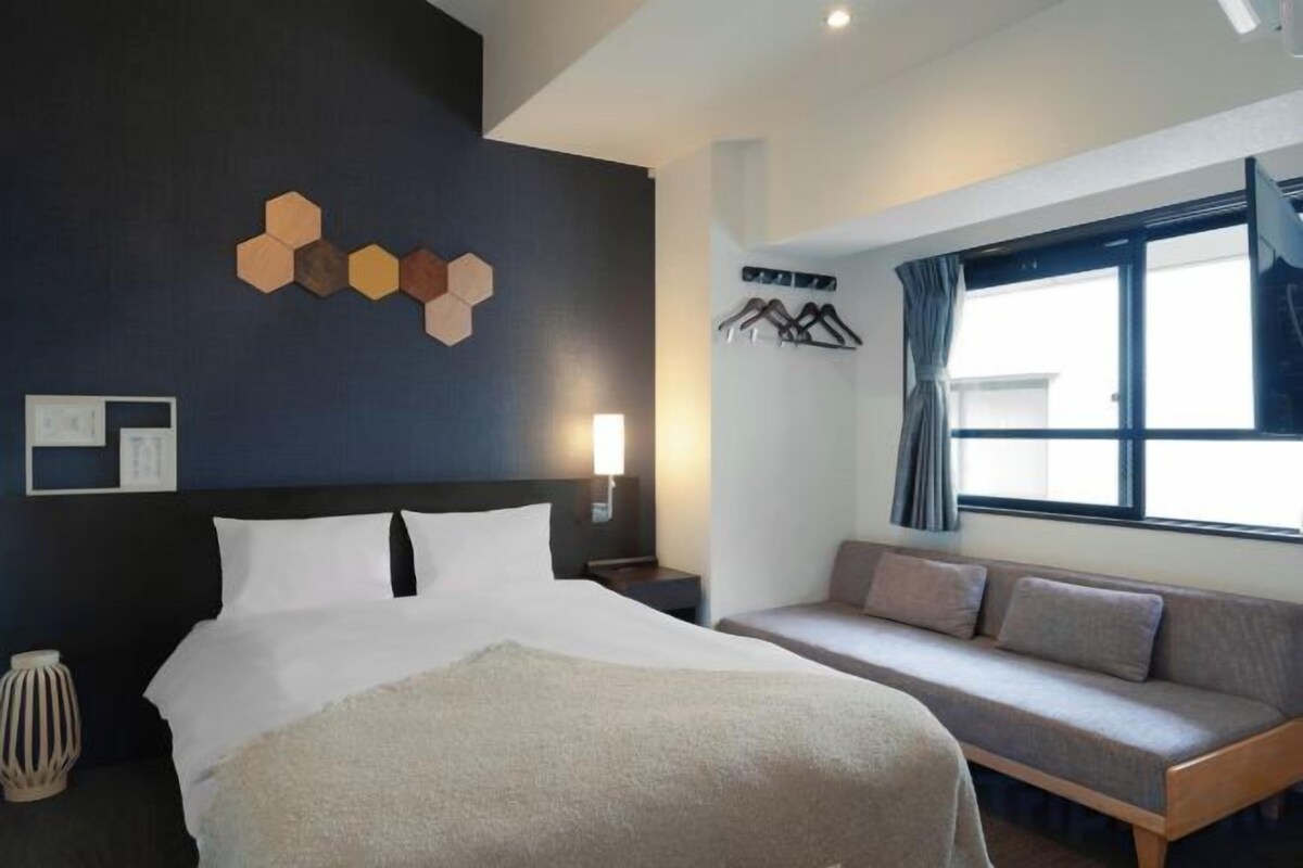 A hotel room with a bed, a sofa, a wall-mounted light, and a window with curtains.