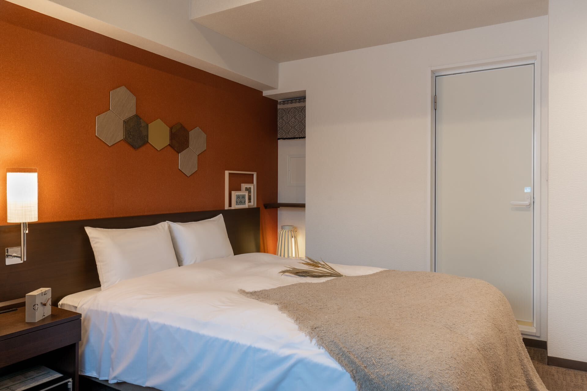 A hotel room with a bed, a nightstand, a wall clock, a lamp, and a door.
