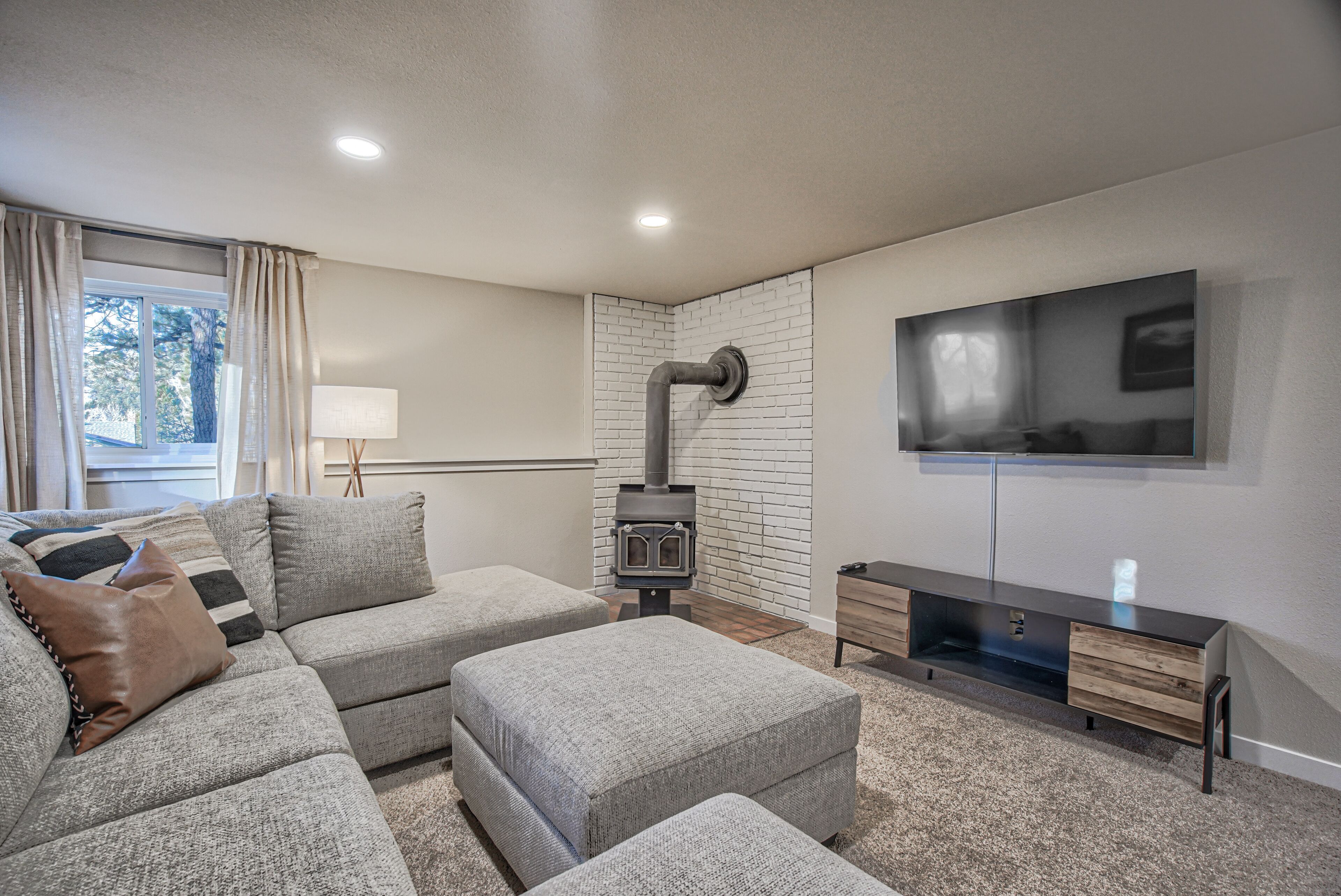 A modern living room with a sectional sofa, ottomans, a TV wall unit, and a wood-burning stove.
