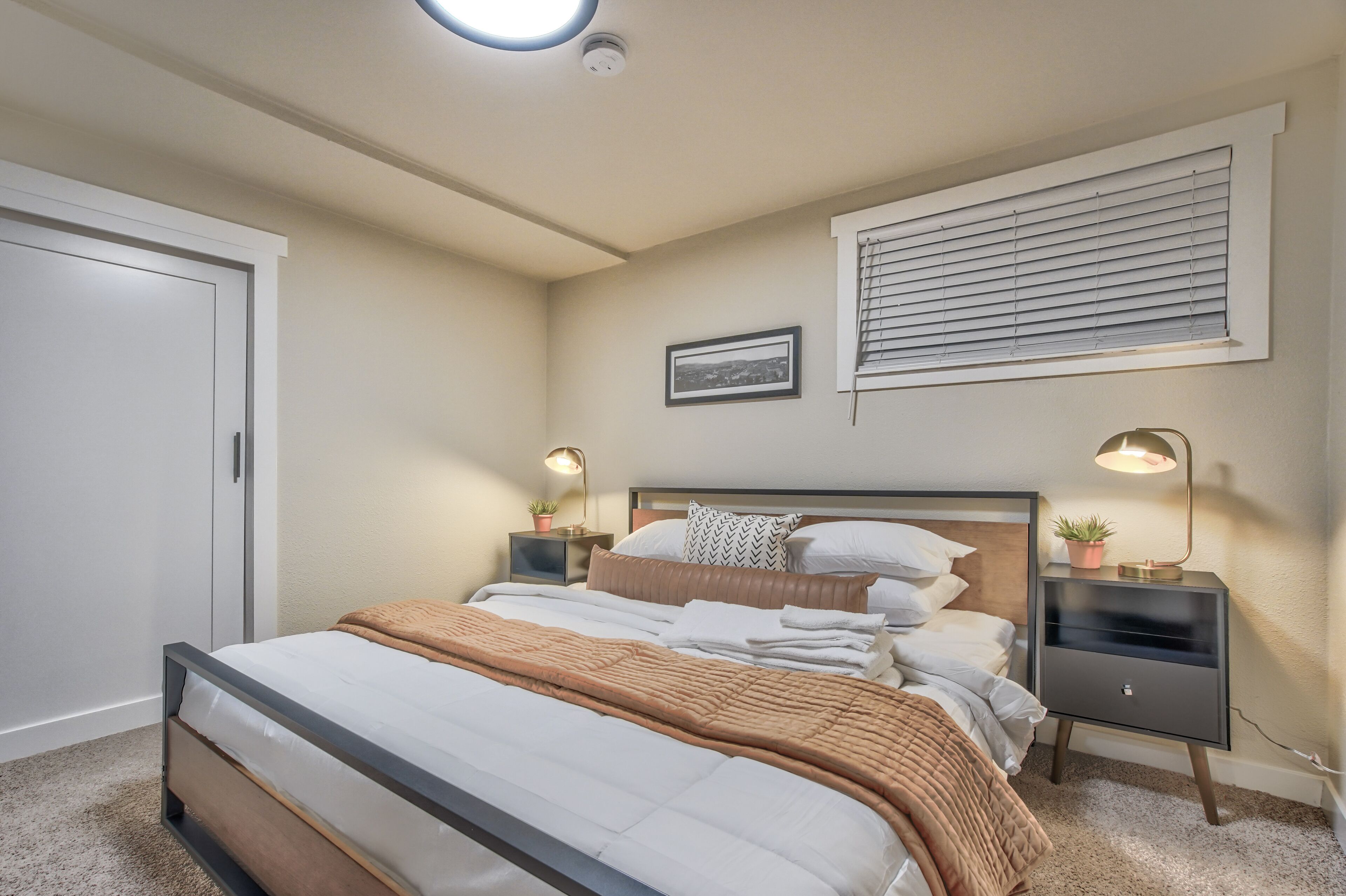 A bedroom with a bed, bedside tables, a nightstand, a wall-mounted picture, and a window with blinds.