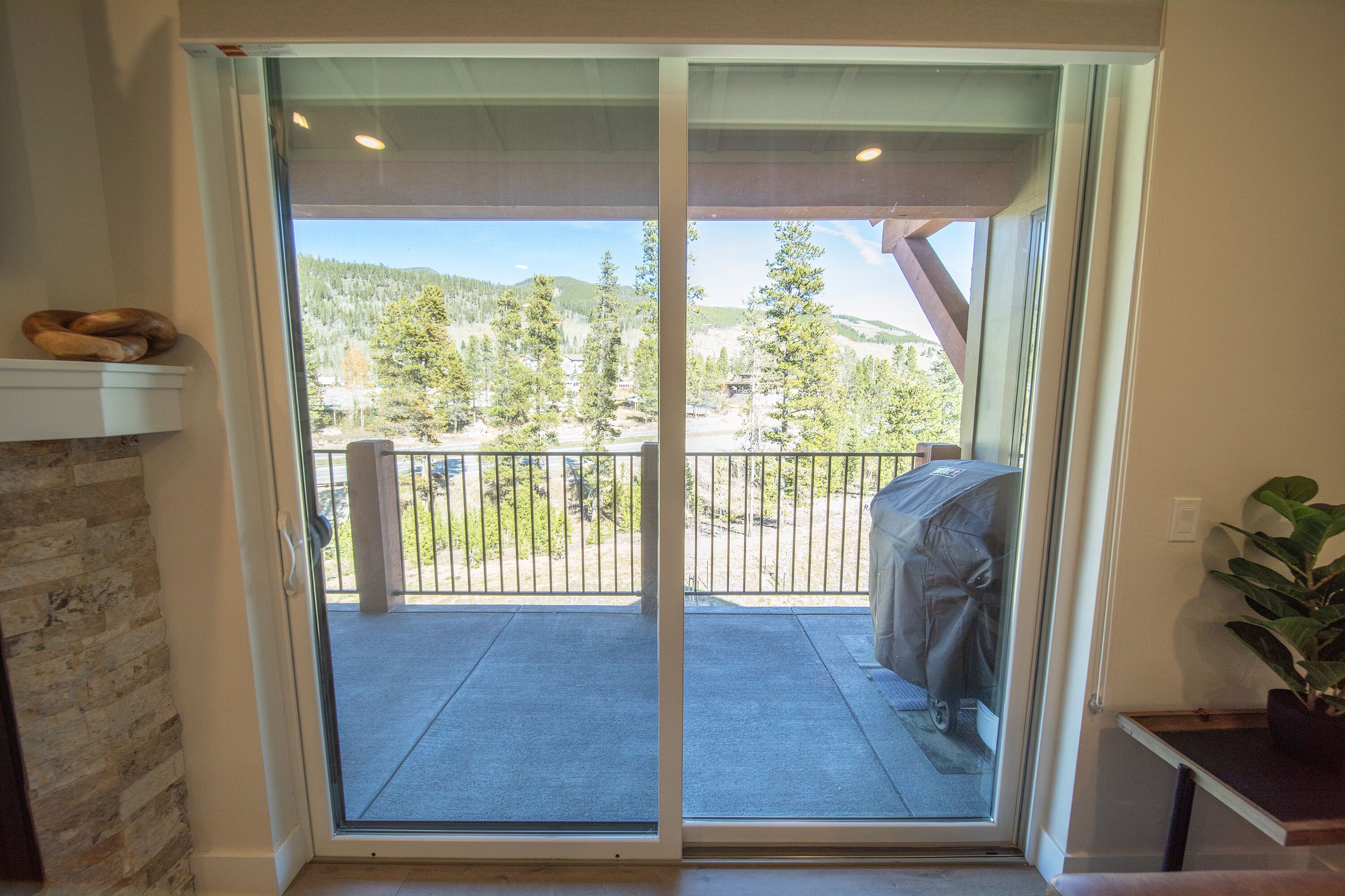 A sliding glass door leading to a balcony with a barbecue grill, a potted plant, and a view of a forested area.