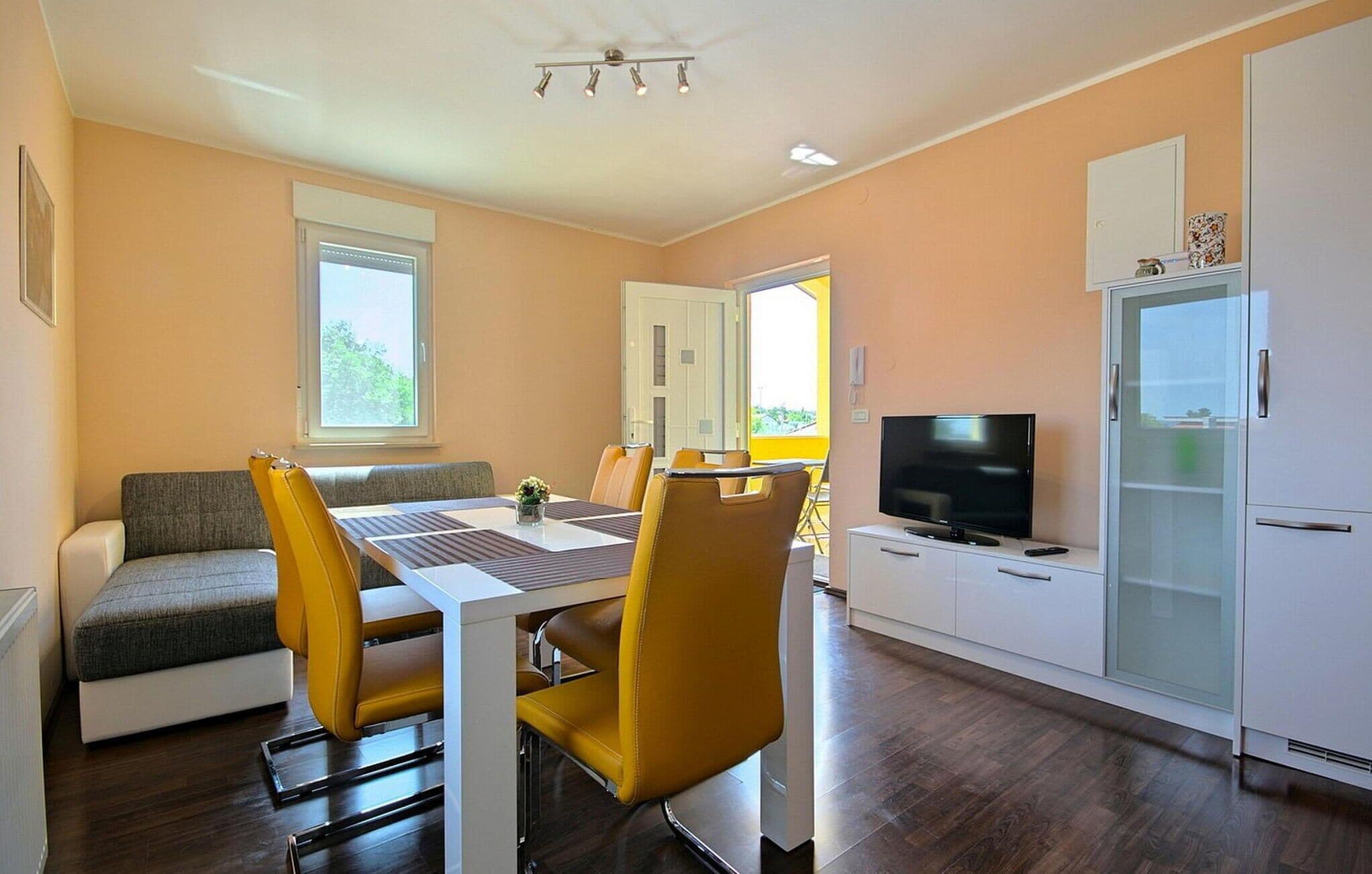 Een moderne woonkamer met een bank, eettafel en stoelen. Er is een televisie en een koelkast.
