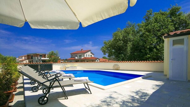 Ein Schwimmbecken mit Liegestühlen und Sonnenschirmen, umgeben von einer gefliesten Terrasse und Blick auf Häuser.