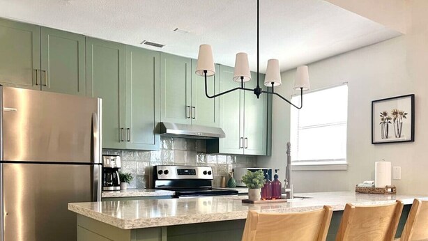Una cocina moderna con armarios verdes, electrodomésticos de acero inoxidable, una isla central con sillas y una lámpara de araña.