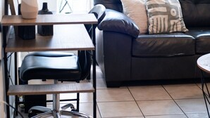 Una sala moderna con un sofá de cuero negro, una mesa de centro de madera y una silla de oficina negra. Hay una planta en maceta sobre una repisa de madera, y una ventana con cortinas blancas.