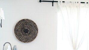 A modern living room with a kitchen area, featuring a round woven wall art, a sofa with cushions, and a dining table with chairs. - 202, Ocean Side, Patio, Laundry, Desk (South Padre Island)
