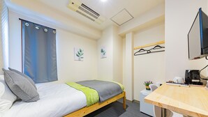 Una cama individual con estructura de madera, una mesita de noche con una planta, un escritorio con una computadora y un televisor instalado en la pared.