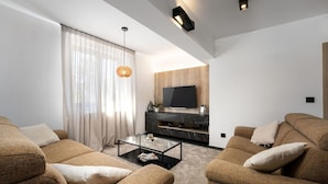 Una sala de estar moderna con un televisor de pantalla plana montado en una pared de madera, dos sofás con cojines, una mesa de centro de vidrio y un ventanal con cortinas translúcidas.