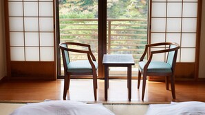 Una habitación con dos sillas de madera, una mesita pequeña, un futón de tatami y vistas a un bosque a través de un celosía japonesa.